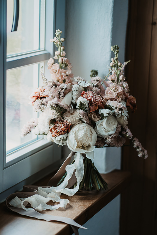 Brautstrauß mit Pfingstrosen, Nelken und Rosen liegt auf einer Fensterbank im hellen Raum der Maisenburg. Natürliches Licht betont die zarten Farben. Aufnahme von Patricia Hamann, Hochzeitsfotografin Ulm.