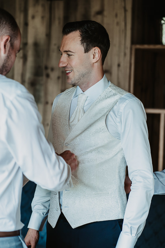 Seitenprofil des Bräutigams wie er von seinem Trauzeugen die Weste zugeknöpft bekommt.