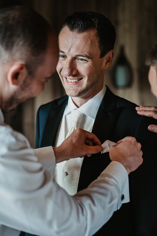 Der Bräutigam lächelt, während sein Trauzeuge ihm an seinem Hochzeitstag das Einstecktuch zurecht macht - Hochzeitsfotograf Rufana Alp
