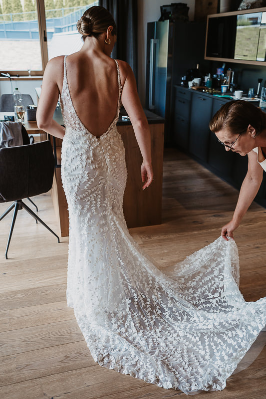 Die Braut steht im Raum eines Chalet, während die Mutter die Schleppe ihres Hochzeitskleids ausbreitet - Hochzeit Rufana Alp