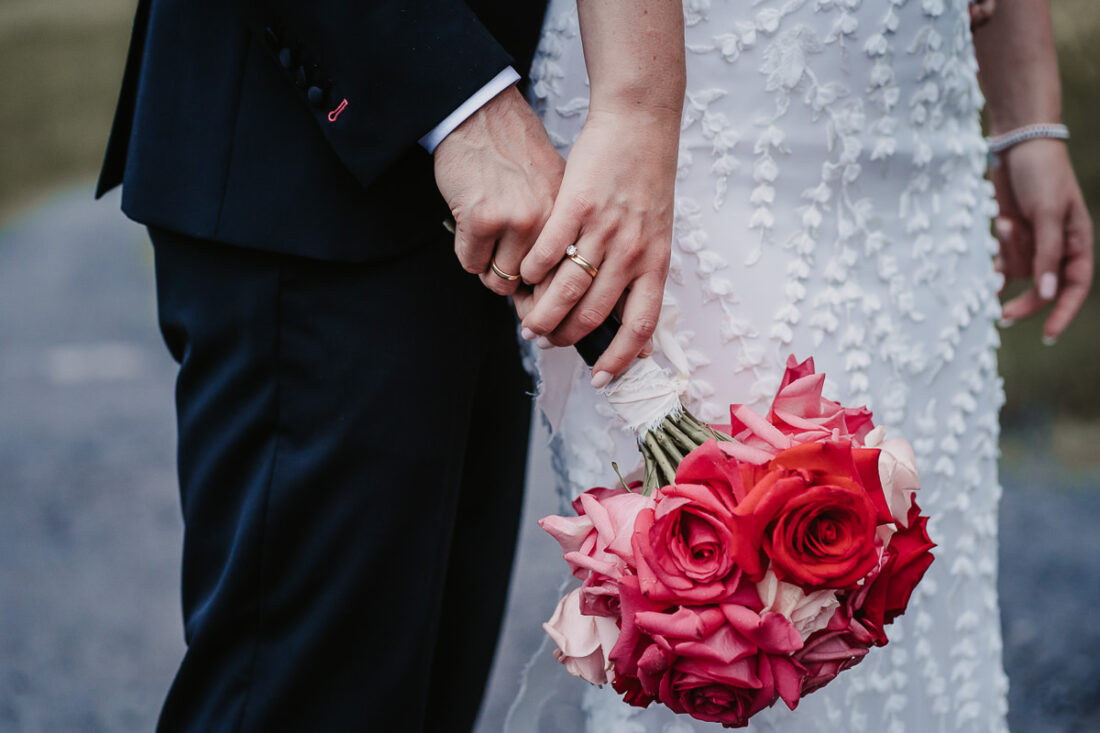 Detailaufnahme von Braut und Bräutigam, die Händchen halten, mit Eheringen und Brautstrauß aus roten Rosen – Hochzeitsfotografie Rufana Alp Tirol