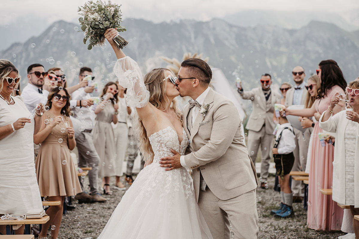 Küssendes Brautpaar wird beim Verlassen ihrer freien Trauung von den Gästen umjubelt. Im Hintergrund sieht man die Berge der österreichischen Alpen.