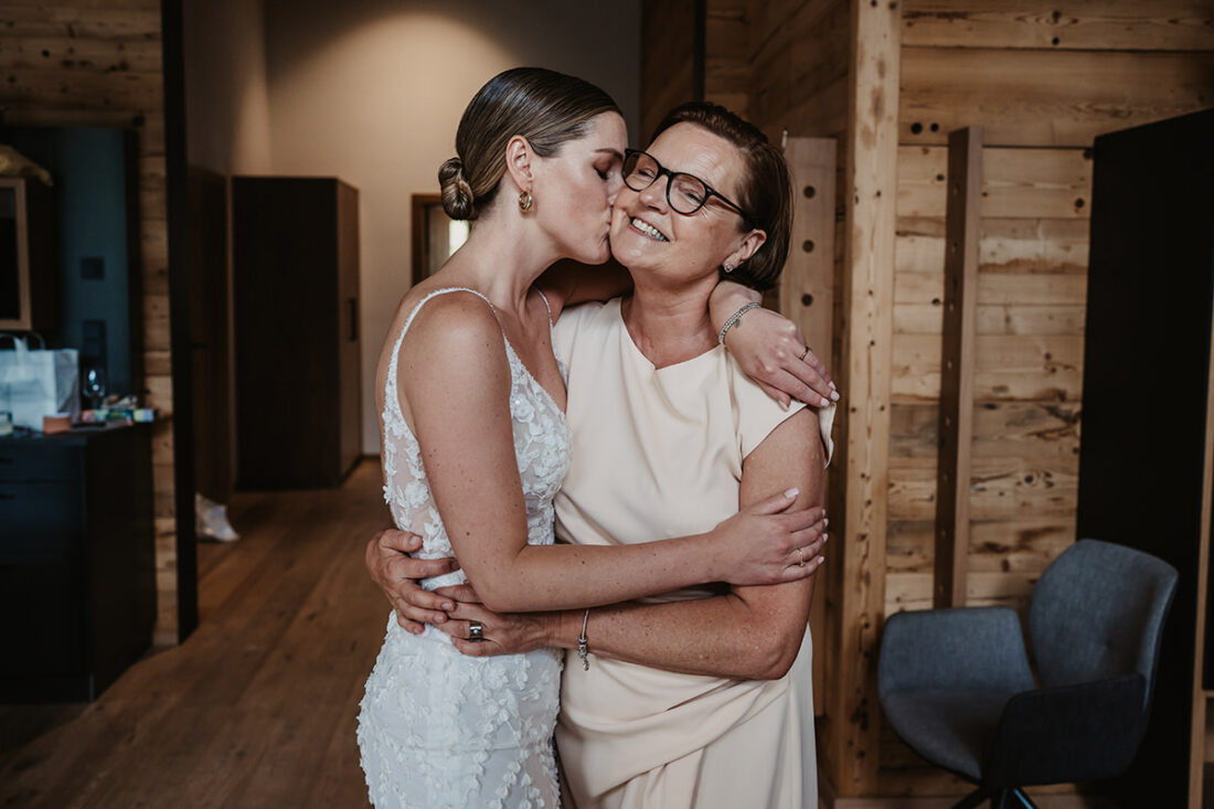 Einem Chalet in den Bergen umarmt die Braut ihre Mutter und gibt ihr einen Kuss auf die Backe - Hochzeitsfotograf Patricia Hamman