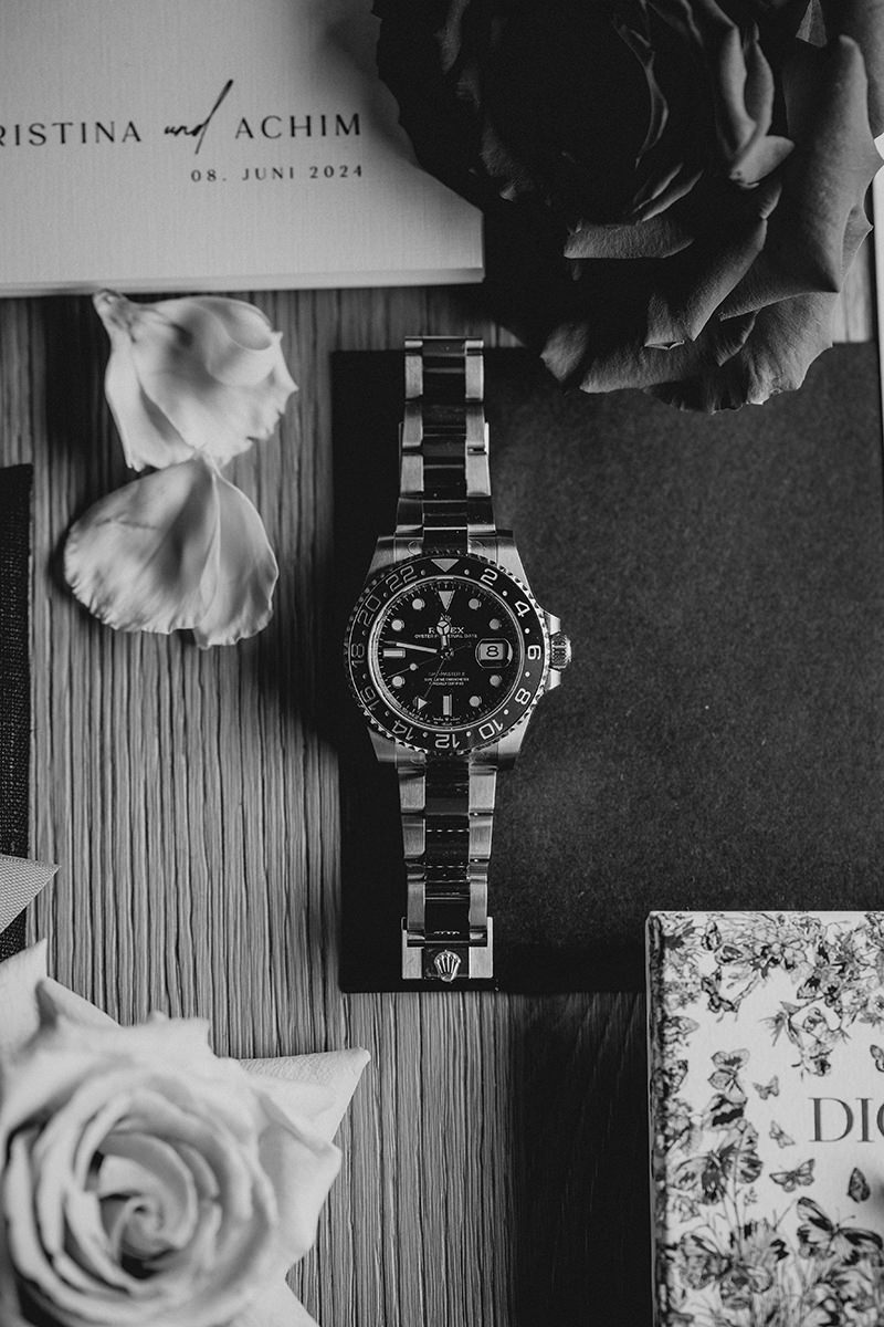 Rolex Uhr liegt in Mitten von Schmuck, Blütenblättern und Hochzeitseinladung.