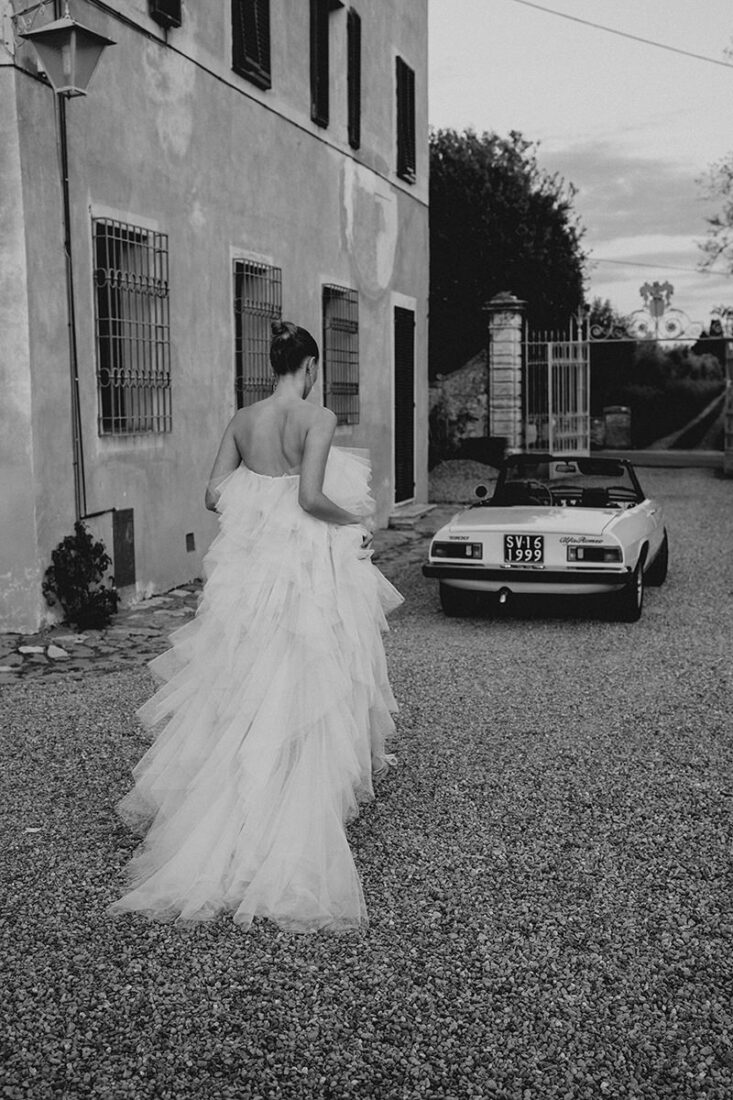 Man sieht die Braut von hinten, wie sie ihr Brautkleid hält und zu einem Oldtimer Alfa Romeo läuft an ihrer Hochzeit in der Toskana.