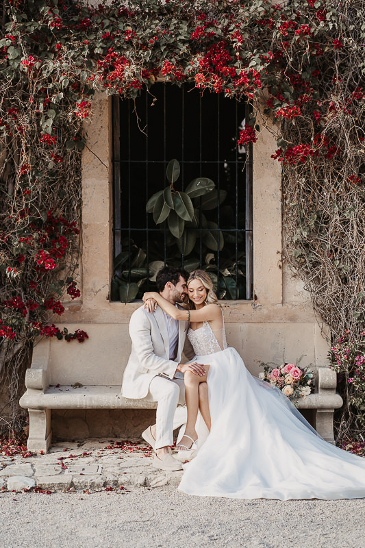 Umarmendes Brautpaar sitzt auf einer Steinbank mit einem Rosenbogen an der Hauswand - Hochzeitsfotograf Patricia Hamann