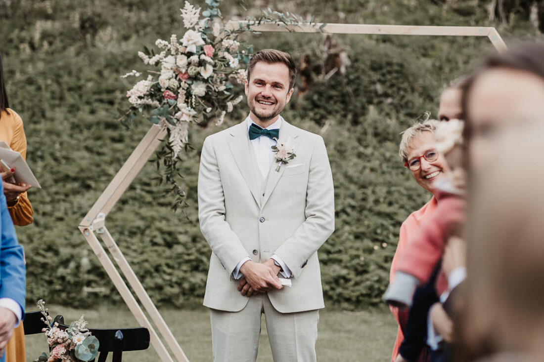 Bräutigam steht während der Trauung auf der Maisenburg und lächelt erwartungsvoll. Die natürliche Kulisse und Blumendeko im Hintergrund unterstreichen die emotionale Atmosphäre. Foto von Patricia Hamann, Hochzeitsfotografin aus Ulm.
