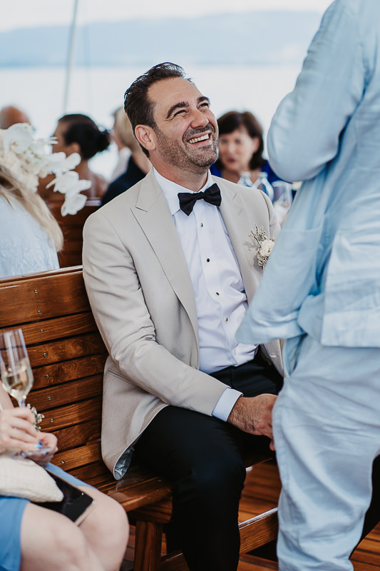 Bräutigam sitzt lachend auf einer Holzbank an Deck der Hohentwiel während der Hochzeit auf dem Bodensee, fotografiert von Patricia Hamann, Hochzeitsfotograf Werft1919