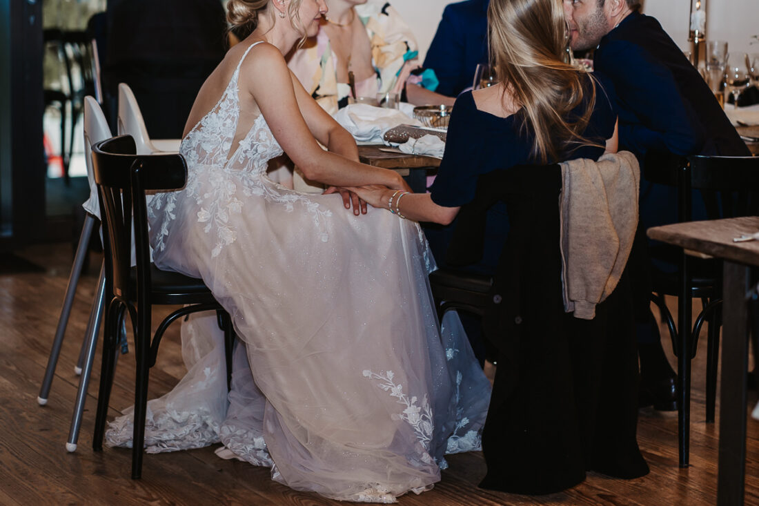 Braut im Spitzenkleid sitzt beim Hochzeitsdinner auf der Maisenburg bei Ulm und hält liebevoll die Hand einer Freundin – fotografiert von Patricia Hamann Hochzeitsfotograf Maisenburg