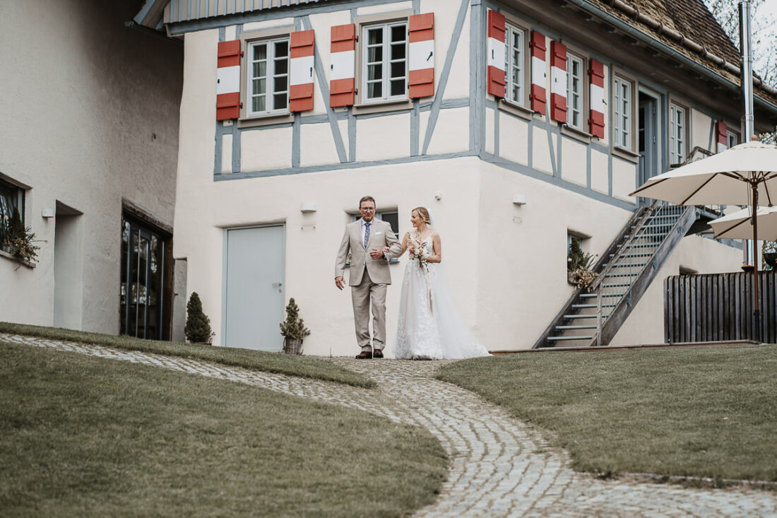 Die Braut wird von ihrem Vater zur freien Trauung auf der Maisenburg geführt. Im Hintergrund das charakteristische Fachwerkhaus des Hofguts. Aufnahme von Patricia Hamann, Hochzeitsfotografin aus Ulm.