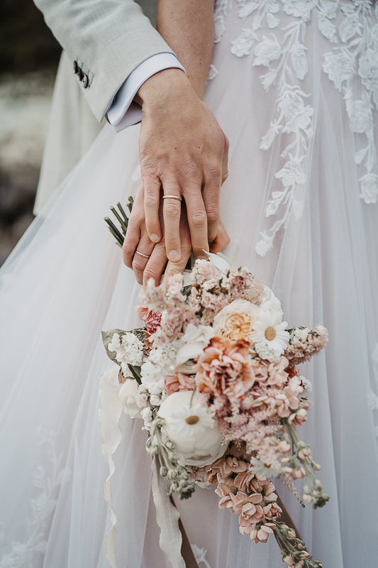 Die Hände von Braut und Bräutigam ruhen liebevoll aufeinander, während der Brautstrauß in Pastelltönen zart herabhängt – fotografiert auf der Maisenburg bei Ulm von Hochzeitsfotografin Patricia Hamann.