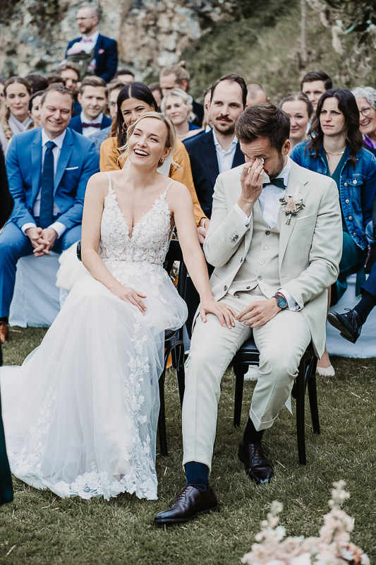 Braut und Bräutigam sitzen während der Trauung auf der Maisenburg. Sie lacht herzlich, während er sich Tränen der Rührung wischt. Foto von Patricia Hamann, Hochzeitsfotografin aus Ulm.