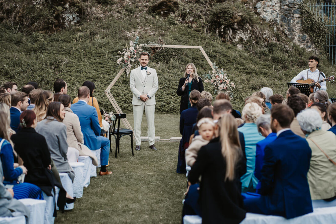 Der Bräutigam steht bei der freien Trauung auf der Maisenburg unter einem Blumenbogen. Eine Rednerin spricht zu den Gästen, während Musiker mit Gitarre begleitet. Hochzeitsreportage von Patricia Hamann, Hochzeitsfotografin aus Ulm.