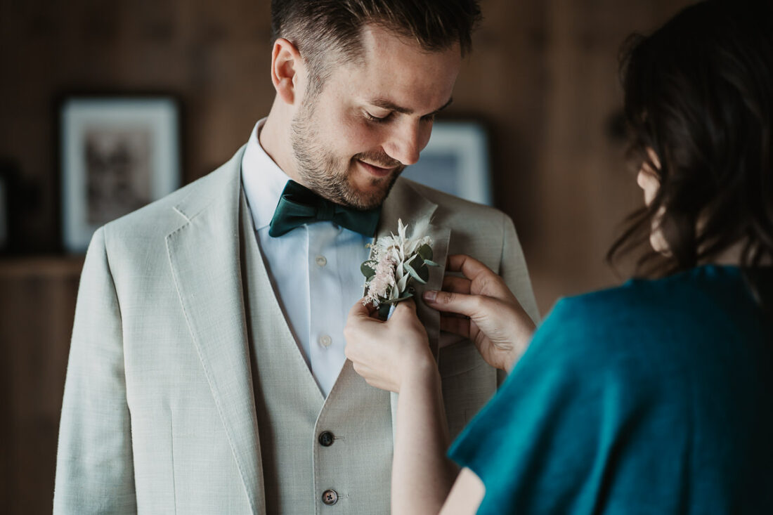Ein Bräutigam im hellen Anzug lächelt, während ihm die Ansteckblume an das Revers gesteckt wird. Aufnahme auf der Maisenburg von Patricia Hamann, Hochzeitsfotografin Ulm.