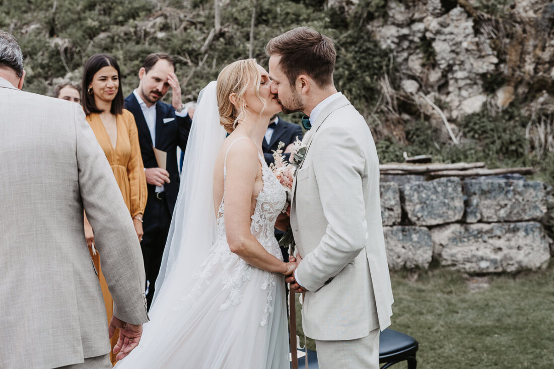 Braut und Bräutigam küssen sich während der Trauung auf der Maisenburg. Im Hintergrund lächeln die Gäste. Aufnahme von Patricia Hamann, Hochzeitsfotografin aus Ulm.