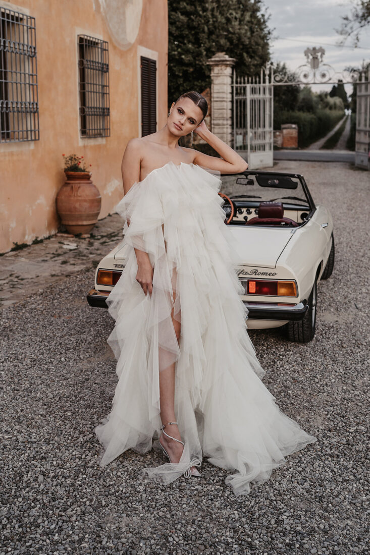 Braut in einem modernen, voluminösen Tüllkleid steht vor einem weißen Cabrio, aufgenommen an einer mediterranen Location auf Ibiza.