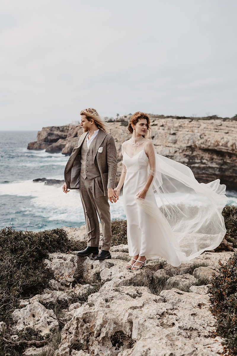 Braut und Bräutigam auf Felsen an der Küste Ibizas, mit wehenden Haaren und Schleier im Wind.