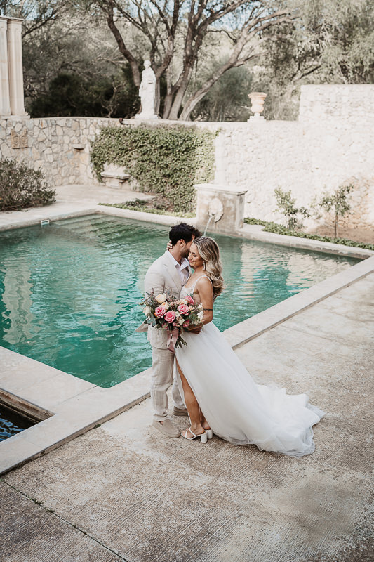 Braut und Bräutigam stehen eng umarmt vor einem türkisfarbenen Pool in einer mediterranen Hochzeitslocation auf Ibiza.