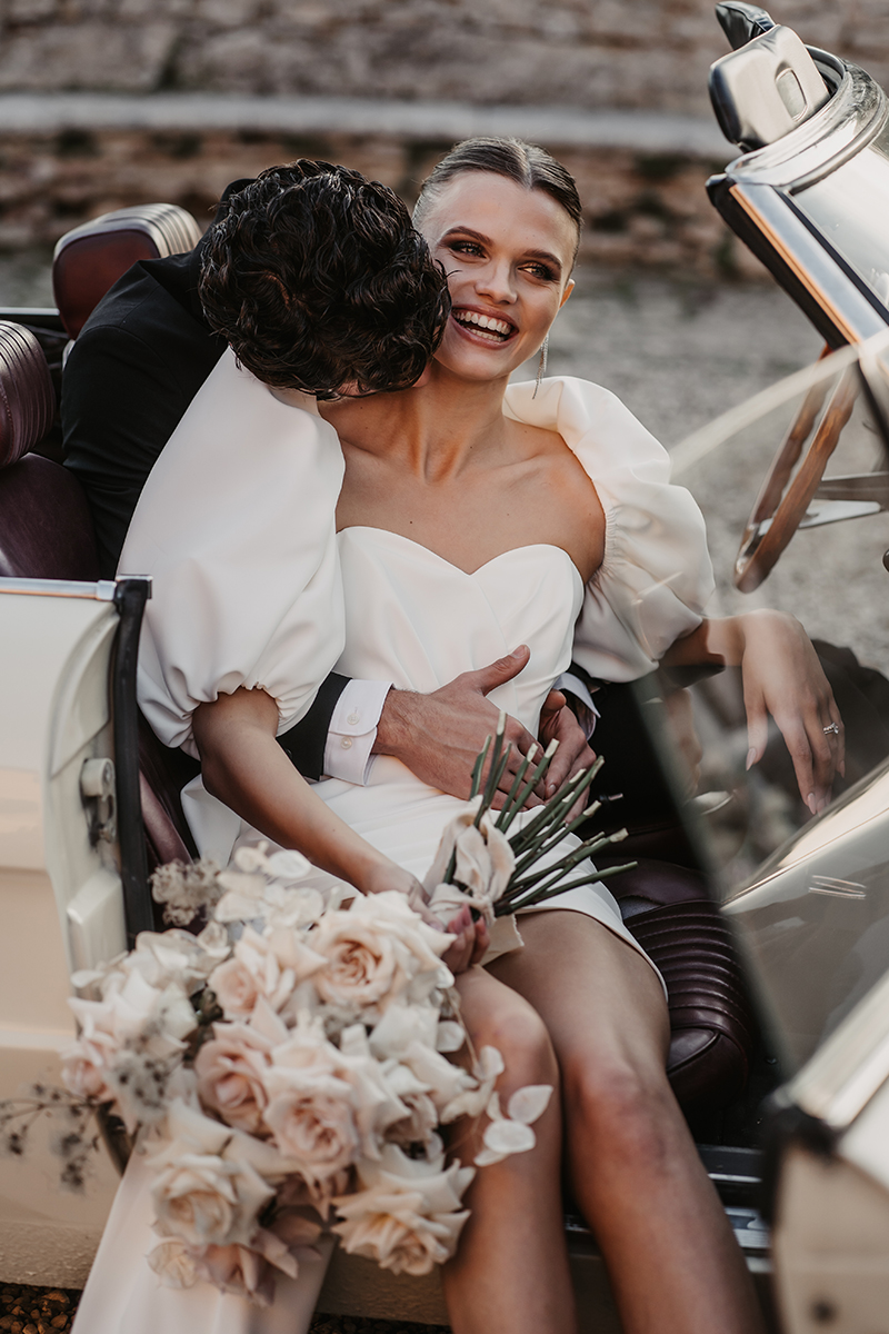 Lachende Braut im Oldtimer, umarmt vom Bräutigam, mit elegantem Brautstrauß auf Ibiza.