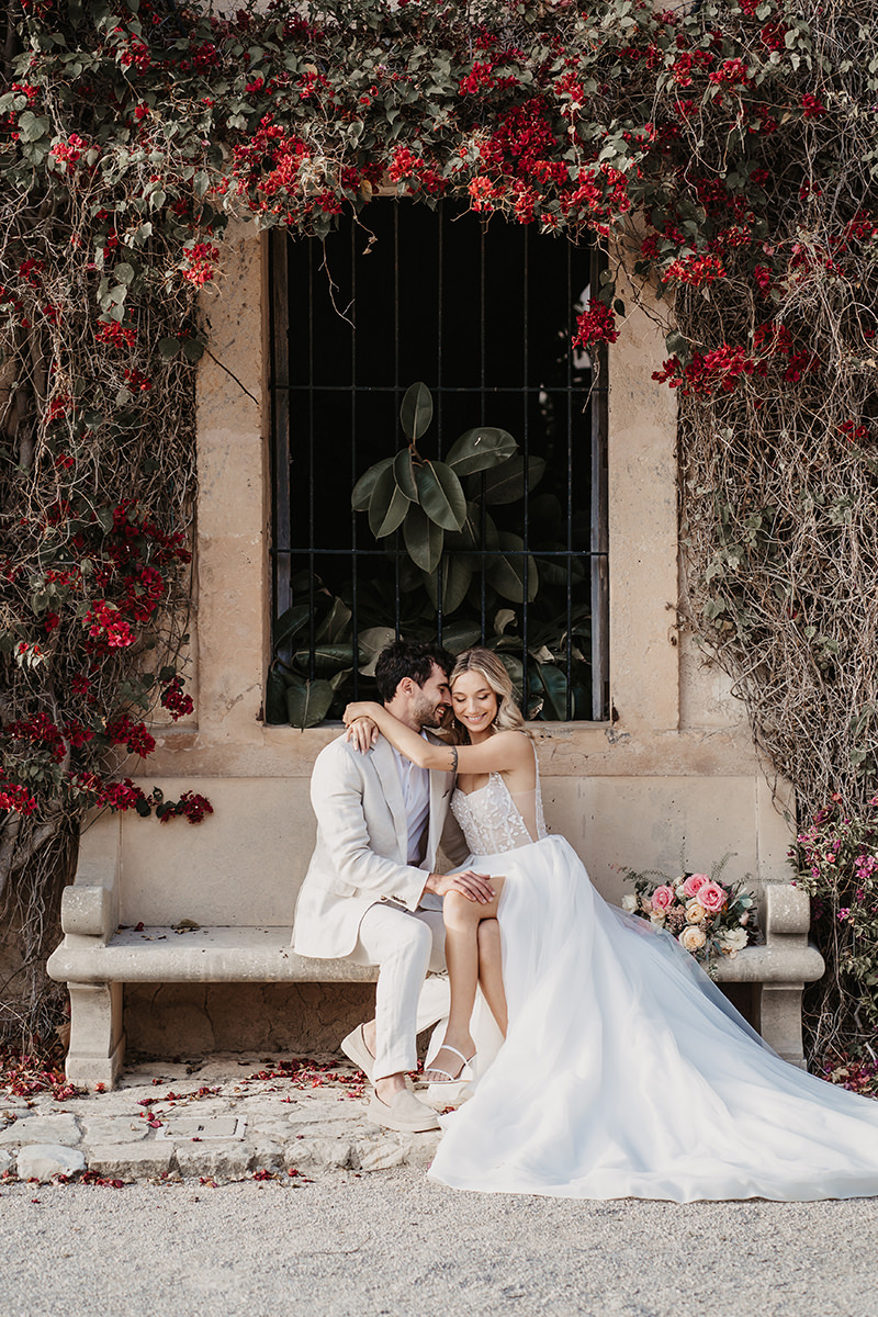 Romantisches Hochzeitspaar auf Ibiza, sitzend auf einer Steinbank unter roten Bougainvillea-Blüten fotografiert von Patricia Hamann.