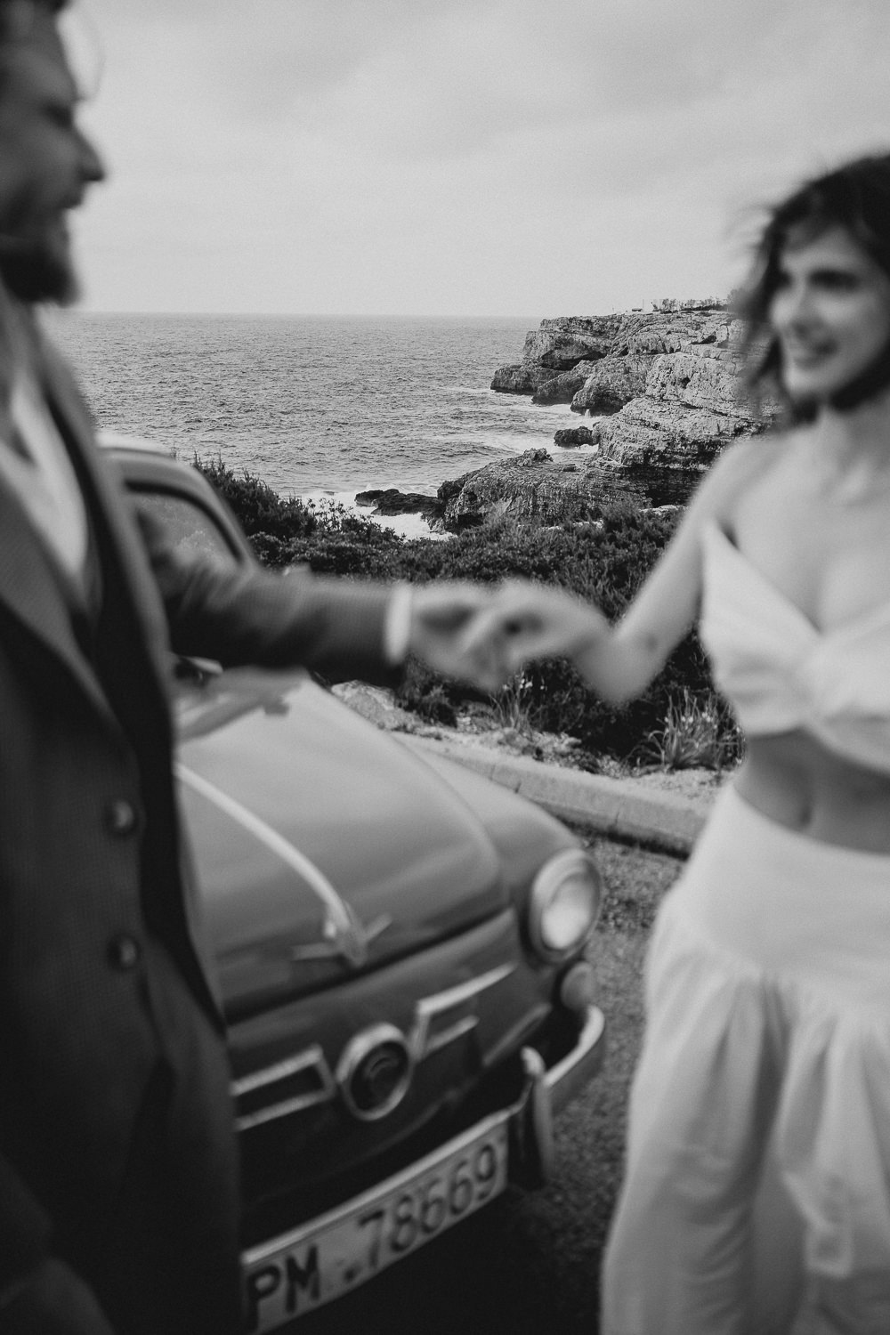 Unscharfe Aufnahme eines Paares, das sich an der Küste Ibizas an den Händen hält, mit Blick auf das Meer und ein klassisches Auto im Hintergrund.