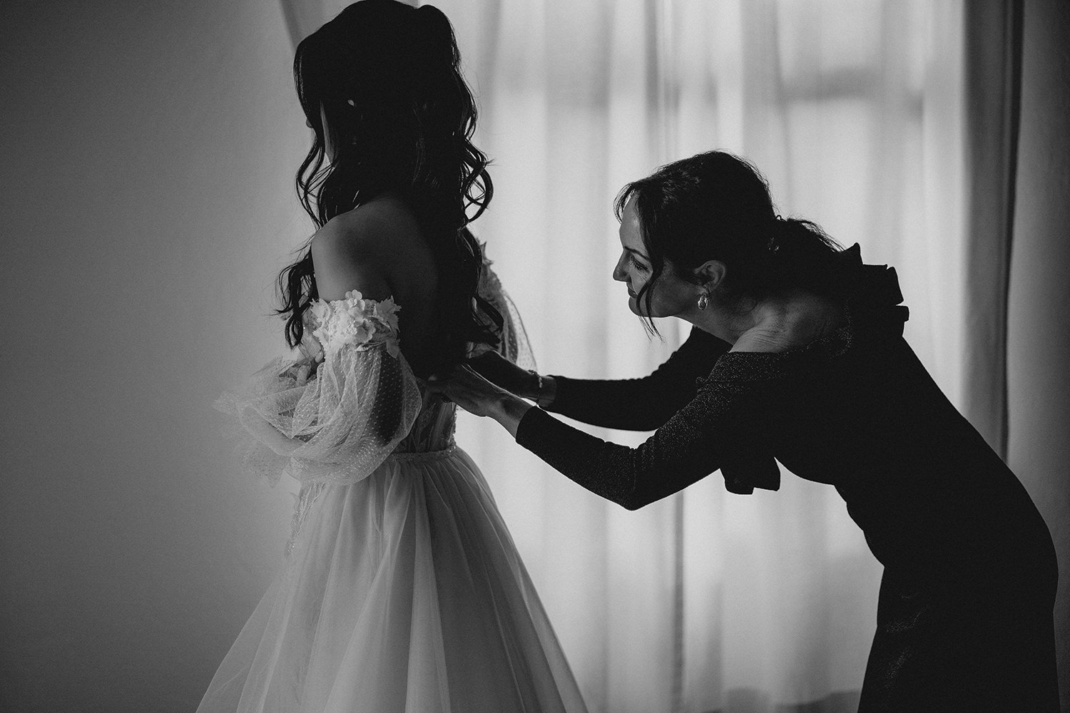 Braut wird von ihrer Mutter beim Getting Ready unterstützt, in dem sie ihr das Kleid am Rücken schließt.