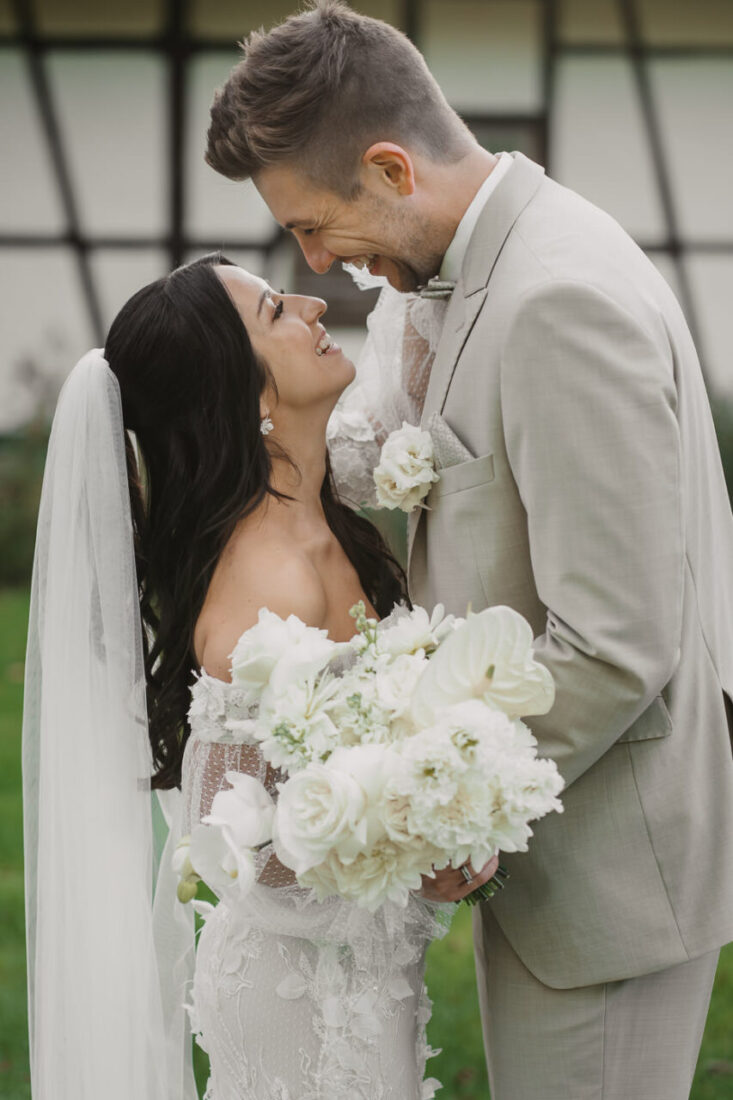 Braut und Bräutigam sehen sich tief in die Augen, während die Braut ihren weißen Brautstrauß in der Hand hält und glücklich lächelt.