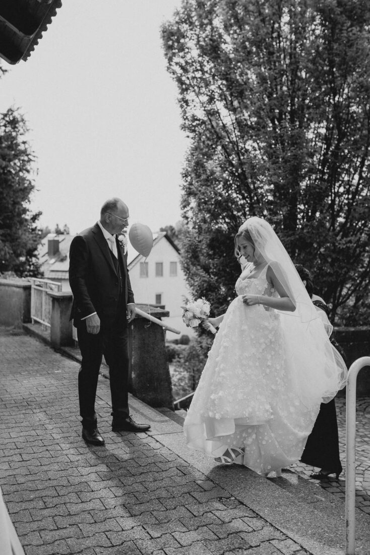 Schwarz-weiß Fotografie einer Braut in einem floralen Hochzeitskleid auf einer Steintreppe vor der Kirche in Lindau
