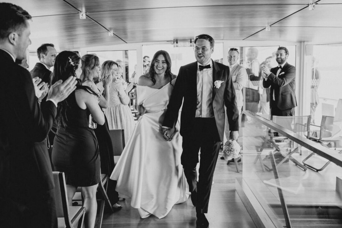 Das strahlende Brautpaar läuft Hand in Hand durch die applaudierenden Gäste im modernen Innenbereich der MS Säntis.
