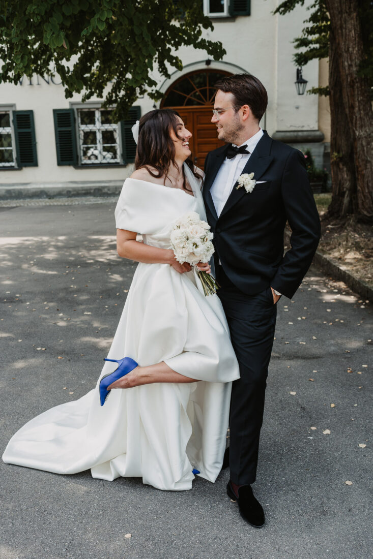 Lachendes Brautpaarportrait vor dem Schloss in Friedrichshafen, Braut zeigt ihre auffälligen blauen Hochzeitsschuhe.