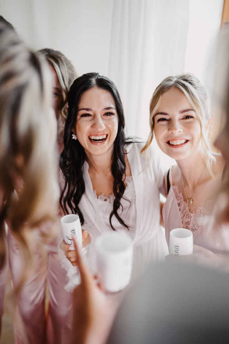 Strahlendes Portrait der Braut, die beim Getting Ready inmitten ihrer Brautjungfern steht und sehr glücklich ist.