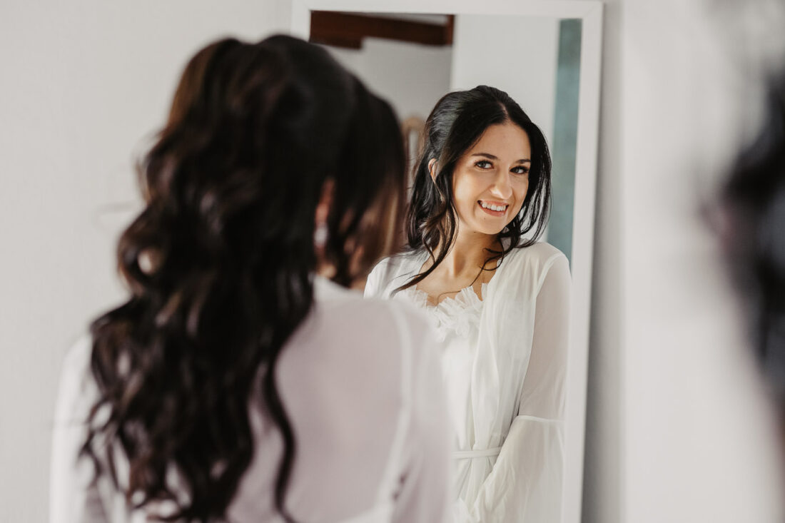Braut betrachtet sich lächelnd im Spiegel beim Getting Ready