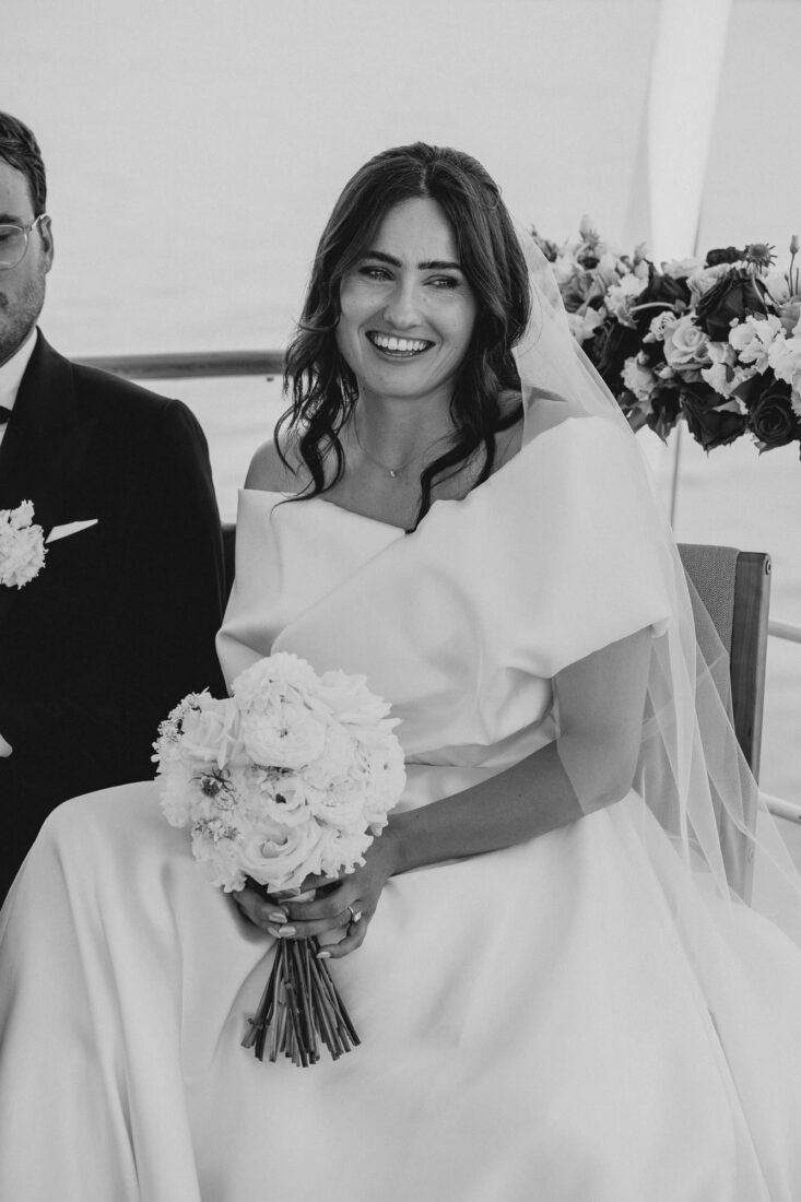 Schwarz-Weiß-Portrait einer lachenden Braut mit Brautstrauß während der Zeremonie auf dem Schiff.