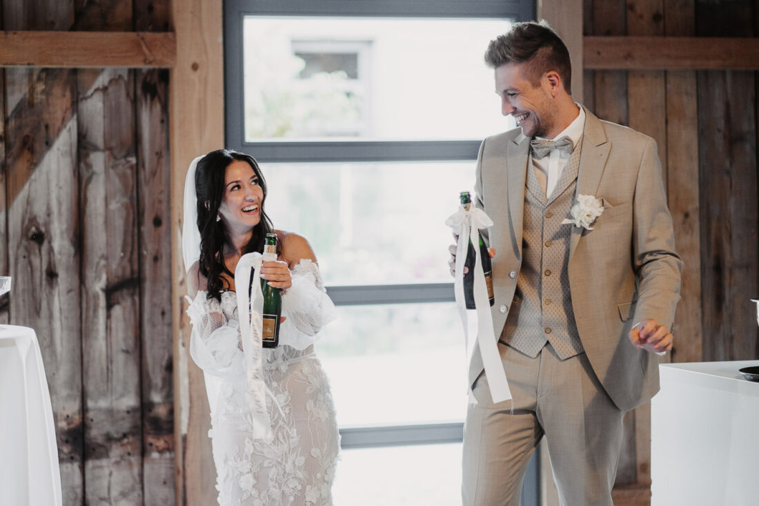 Braut und Bräutigam lachen herzlich, während sie die Champagnerflaschen für die Pyramide öffnen.
