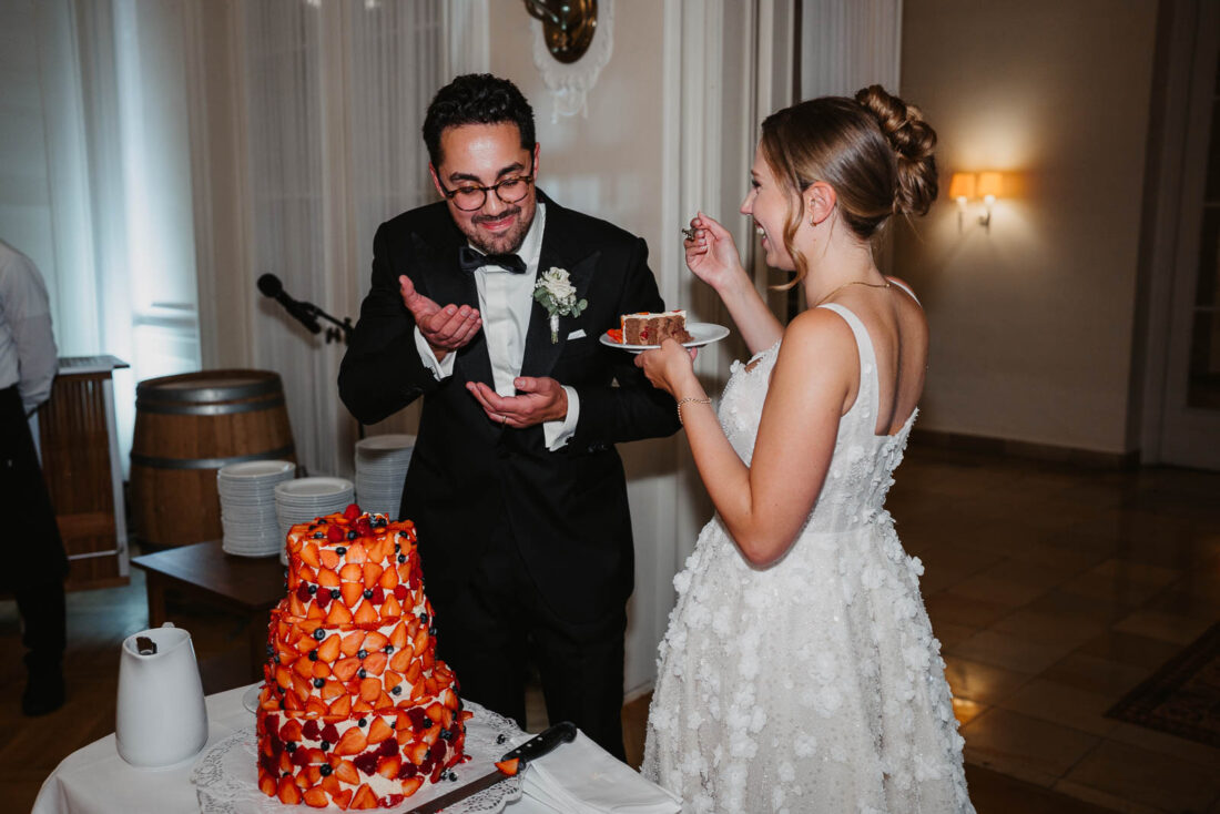 Braut und Bräutigam füttern sich gegenseitig lachend mit einem Stück ihrer Hochzeitstorte.