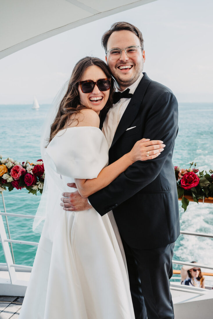Glückliches Brautpaar mit Sonnenbrillen umarmt sich auf dem Deck der MS Säntis vor der Kulisse des Bodensees.