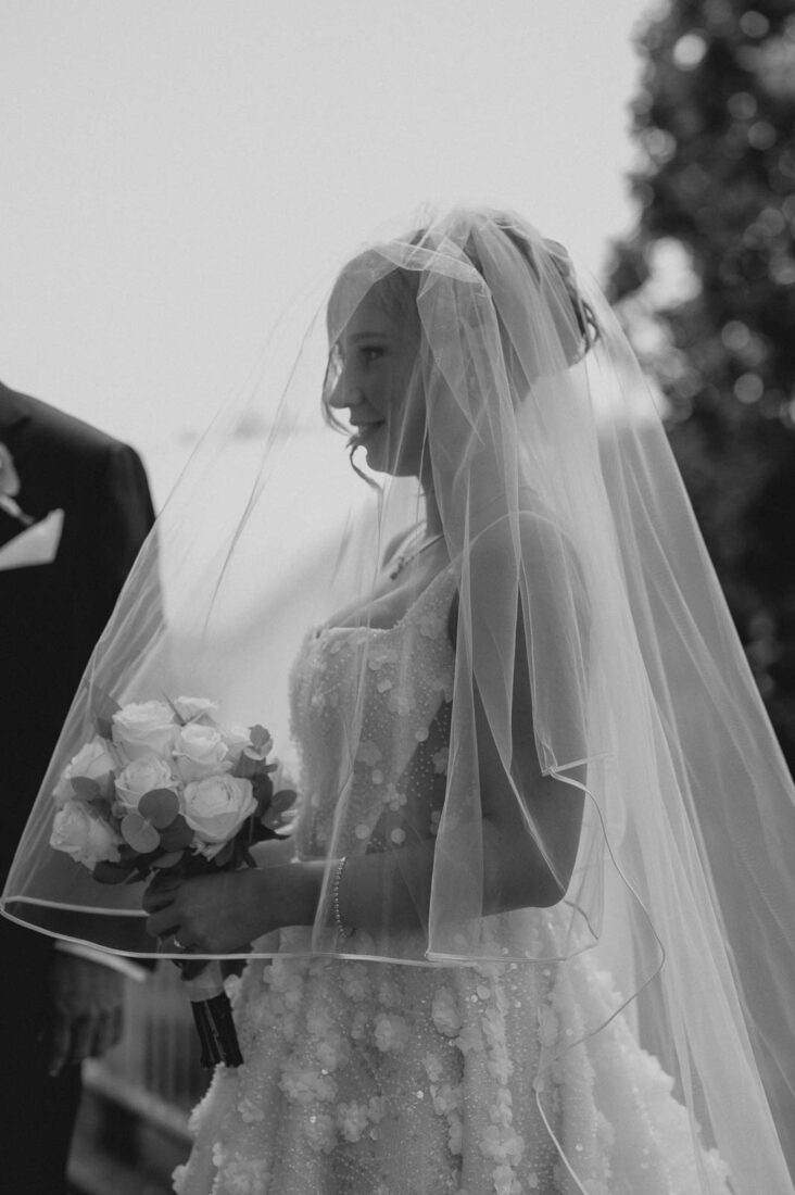 Detailreiches S/W Porträt einer Braut mit langem Schleier und Rosen-Brautstrauß am Bodensee.