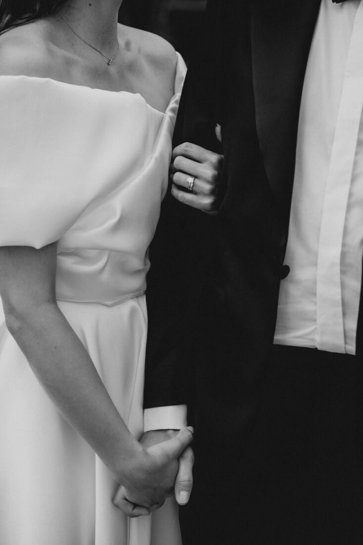 Nahaufnahme der Hände des Brautpaares mit Eheringen vor dem schwarzen Hochzeitsanzug und weißem Kleid.