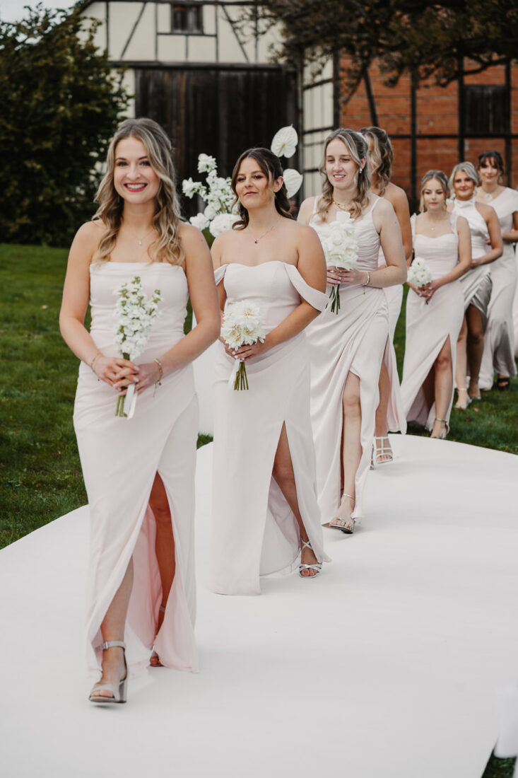 Eine Reihe von Brautjungfern in rosa Kleidern zieht über den weißen Teppich zur freien Trauung auf dem Steinbachhof ein.