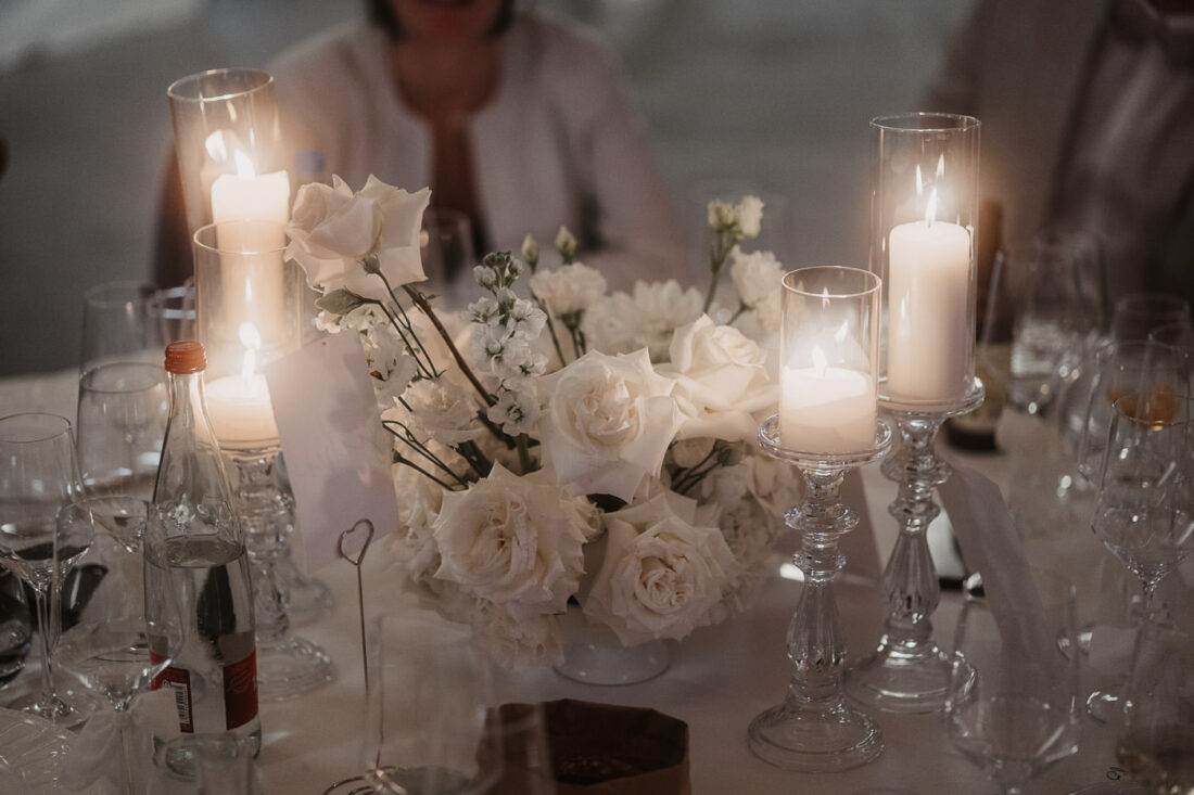 Romantische Tischdekoration mit weißen Rosen, hohen Glasleuchtern und brennenden Kerzen bei einer Hochzeit.