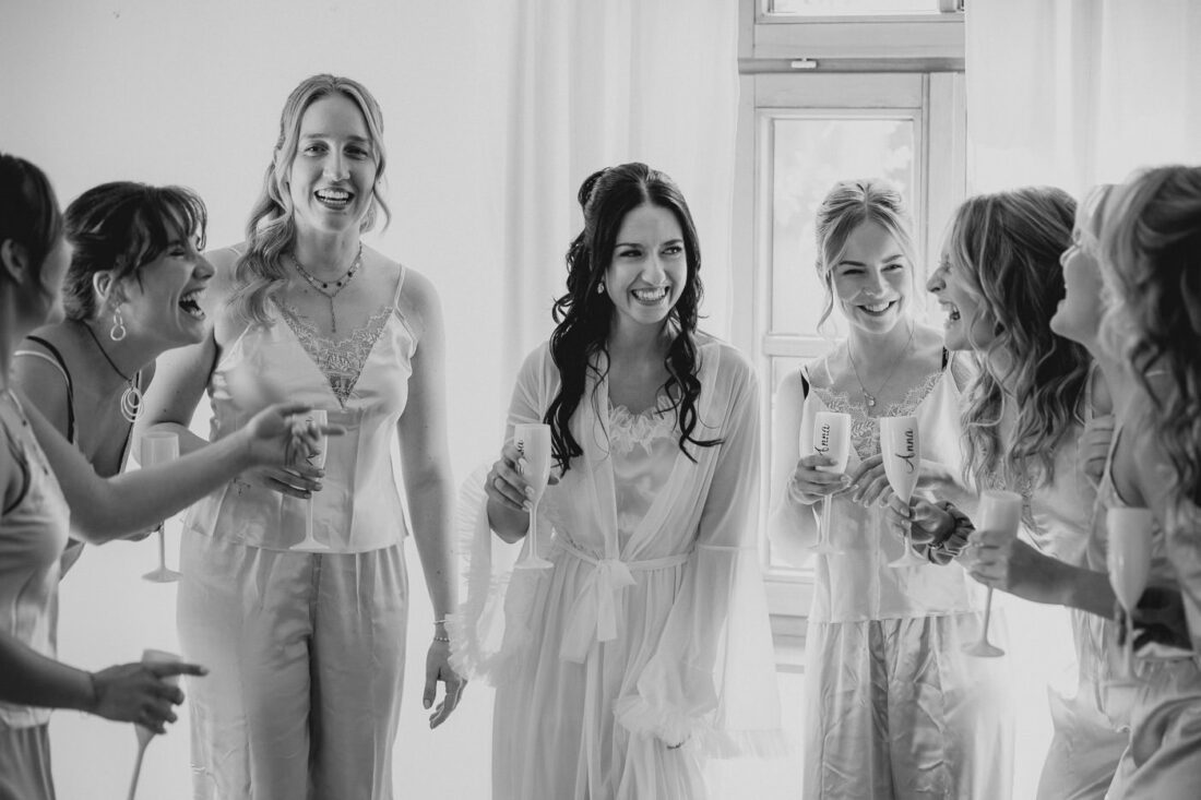 Braut steht lachend zwischen ihren Brautjungfern beim Getting Ready und sie stoßen mit Champagner an.