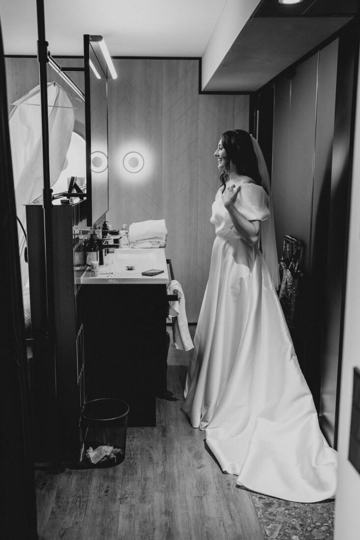 Braut in ihrem eleganten weißen Hochzeitskleid steht lächelnd vor dem Spiegel während des Getting Ready.