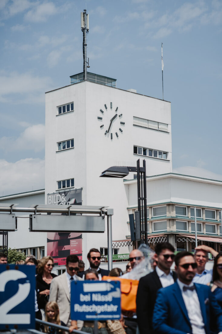 Der weiße Uhrturm am Hafen von Friedrichshafen unter blauem Himmel mit Gästen im Vordergrund.