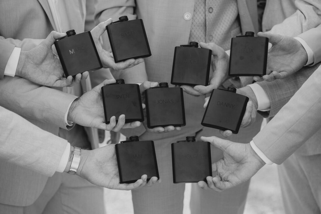 Der Bräutigam und die Trauzeugen halten gravierte Flachmänner in die Kamera.