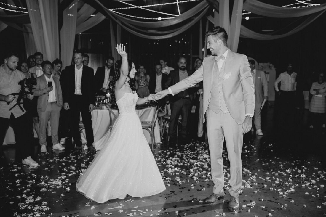 Die Braut streckt beim Hochzeitstanz die Hand die Höhe und lacht ihren Bräutigam verliebt an, während die Gäste im Kreis stehen uns ihnen applaudieren