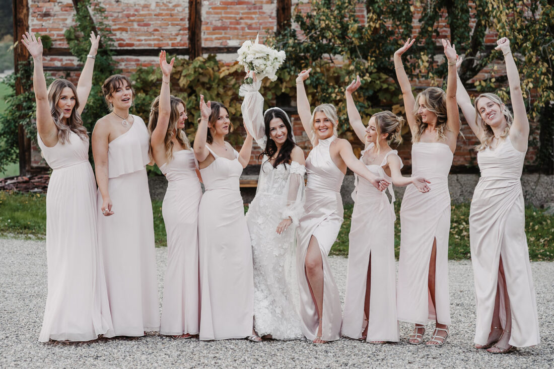Fröhliche Brautgruppe in hellrosa Kleidern jubelt gemeinsam vor einer rustikalen Backsteinmauer am Steinbachhof