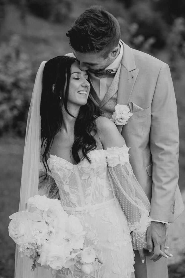 Bräutigam küsst bei den Hochzeitsfotos seine Braut sanft auf die Stirn, während sie glücklich lächelt und seine Hand hält.