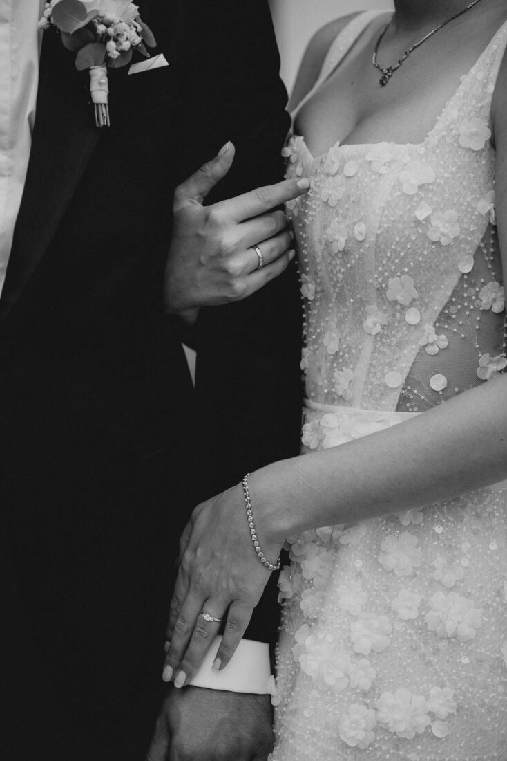 Nahaufnahme der Hände des Brautpaares mit Eheringen und Details des Brautkleids.