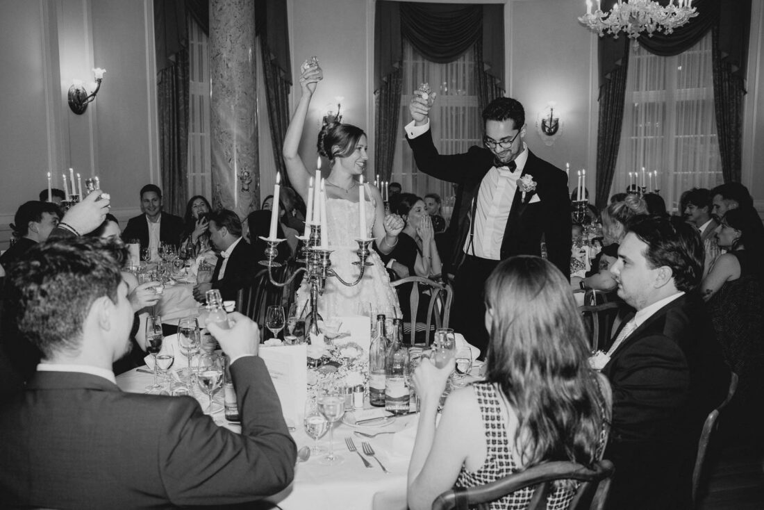 Braut und Bräutigam stehen lachend an ihrem Tisch im Hotel Bad Schachen und stoßen mit ihren Gästen an.