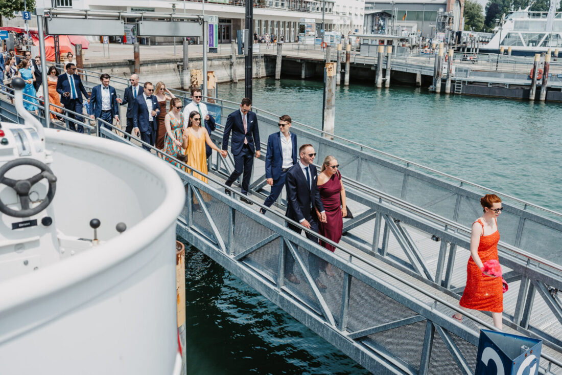 Festlich gekleidete Hochzeitsgäste laufen bei strahlendem Sonnenschein über den Steg auf das Schiff im Hafen von Friedrichshafen.